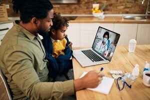 father holding his son on his knees while on a telemedicine call with a doctor
